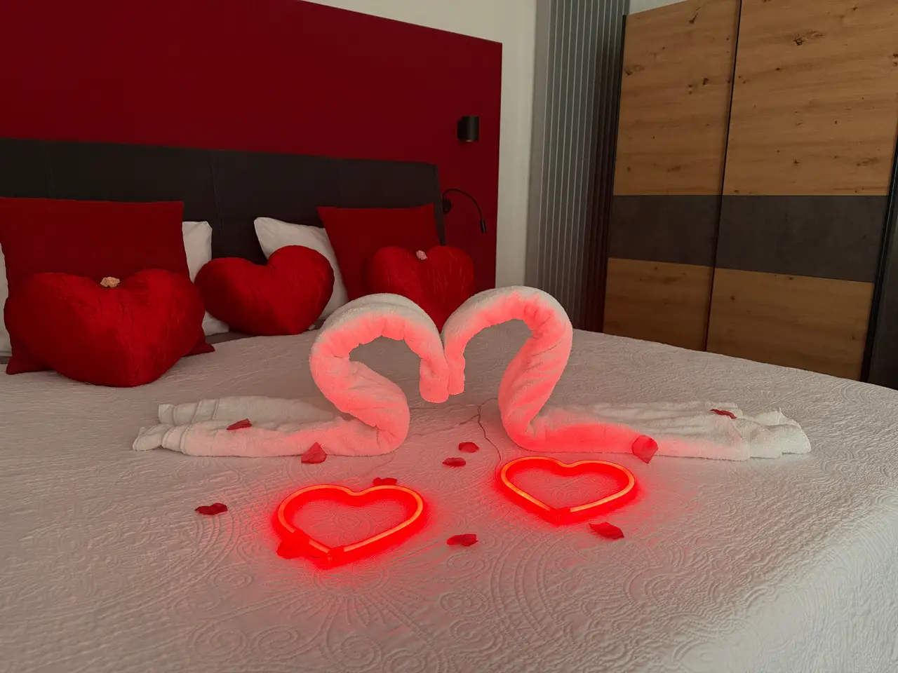 Romantisch dekoriertes Doppelbett in der Romantik-Suite bei Heilbronn mit Handtuch-Schwänen in Herzform, leuchtenden LED-Herzen, roten Herzkissen und Rosenblättern vor stilvoller roter Akzentwand.